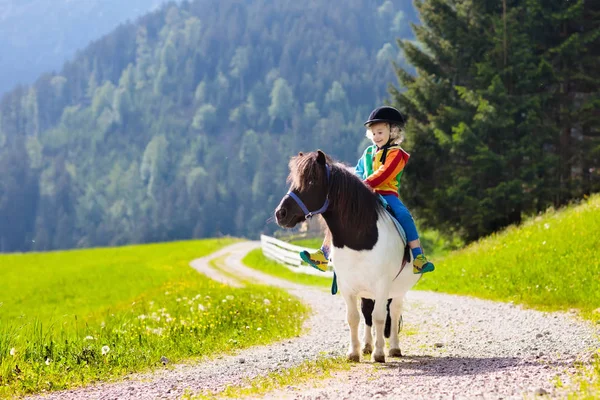 Kids riding pony in the Alps mountains. Family spring vacation on horse ranch in Austria, Tirol. Children ride horses. Kid taking care of animal. Child and pet. Little boy in saddle on pony.