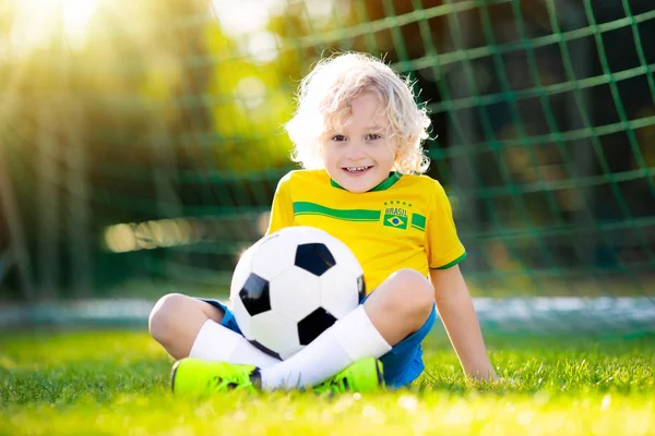 Kids play football on outdoor field. Brazil team fans. Children score a goal at soccer game. Little boy in Brazilian jersey and cleats kicking ball. Football pitch. Sports training for player.