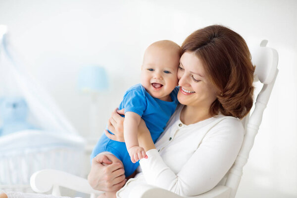 Young mother holding her newborn child. Mom nursing baby. Woman and new born boy in white bedroom with rocking chair and blue crib. Nursery interior. Mother playing with laughing kid. Family at home