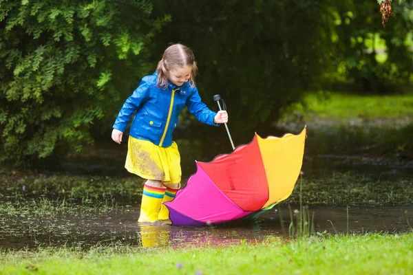 雨の夏の公園で遊ぶ少女 雨の中歩いて泥の水たまりにジャンプ色鮮やかなレインボー傘 青いコートの子 子供秋のシャワーで楽しんで あらゆる天候での野外活動 — ストック写真