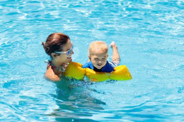 Anne ve bebek tropikal tatil köylerinin havuzunda yüzüyorlar. Çocuk yüzmeyi öğreniyor. Anne ve çocuk suda oynuyorlar. Egzotik bir yerde aile tatili. Çocuklar için aktif ve sağlıklı spor.