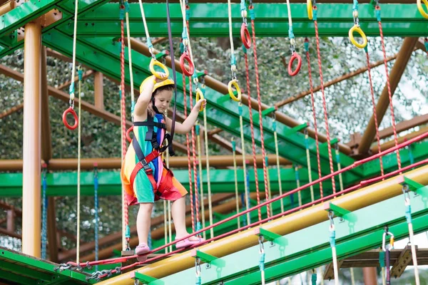 Child in forest adventure park. Kids climb on high rope trail. Agility and climbing outdoor amusement center for children. Little girl playing outdoors. School yard playground with rope way.