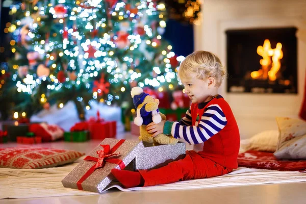 子供は自宅のクリスマス ツリーに存在を開きます 子供へのクリスマス プレゼントやおもちゃ 暖炉のそばで雪フレーク飾り付きニット セーターで小さな男の子 子供を持つ家族は冬の休日を祝うため — ストック写真