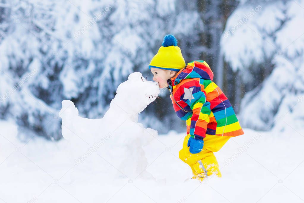 Kid making snowman in snowy winter park. Children play in snow. Baby boy in colorful jacket and hat building snow dinosaur in winter garden after snowfall. Outdoor fun on cold winter day.