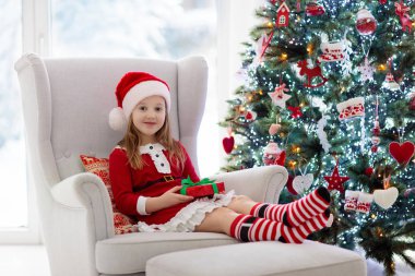 Noel ağacı süsleme ve açma çocuk sunmak ve evde hediye. Xmas hediye ve oyuncaklar ile evlat. Noel Baba elbise ve şapka şömine, küçük kız. Çocuklar ailesiyle kutlamak kış tatili.