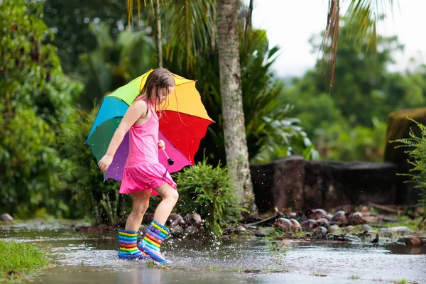 雨の中で遊んでいる子供 傘やレインブーツを着た子供たちは大雨の中屋外で遊ぶ 泥だらけの水たまりに飛び込む少女 子供たちは雨の秋の天気で楽しみます 熱帯低気圧の中を走る子供 — ストック写真