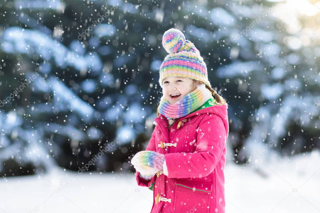 Child playing with snow in winter. Little girl in colorful jacket and knitted hat catching snowflakes in winter park on Christmas. Kids play and jump in snowy forest. Snow ball fight for children.