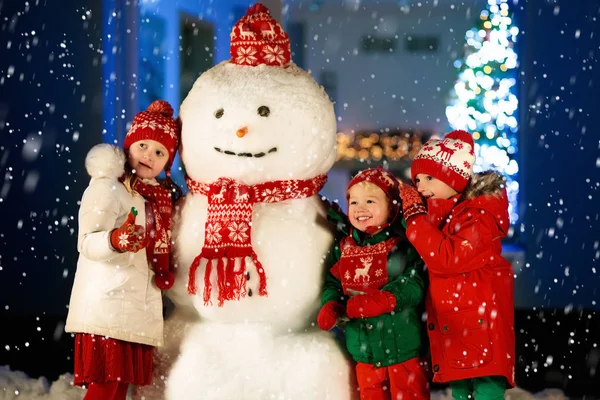 Kids build snowman on Christmas eve. Children in snowy backyard next to window to living room with Christmas tree and decorated fireplace. Boy and girl building snow man on Xmas evening.