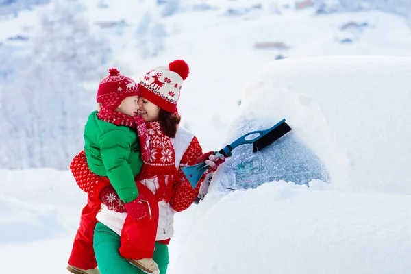 母亲和孩子在暴风雨后刷洗和铲去车上的雪 带着冬刷和刮胡刀的父母和孩子在一夜暴风雪之后清扫了家里的汽车 山里的圣诞假期 — 图库照片