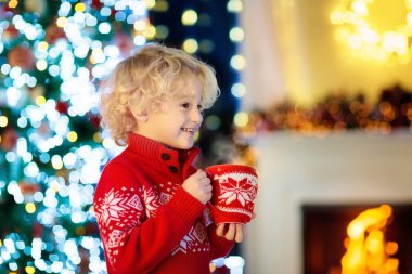 Çocuk evde Noel ağacı sıcak çikolata içme. Süslü içecek kupa. Sıcak kakao kırmızı Kupası tutan Xmas süsleme ile örme kazak içindeki küçük çocuk. Çocuklar ailesiyle kutlamak kış tatili.