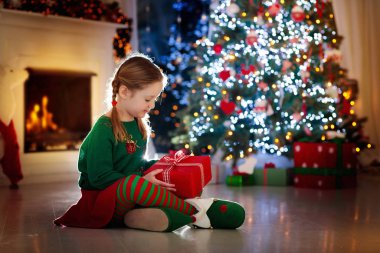 Evdeki Noel ağacında çocuk açılışı hediyesi. Noel hediyeleri ve oyuncakları olan elf kostümlü bir çocuk. Elinde hediye kutusu ve şöminede şekerlemesi olan küçük bir kız. Aile kış tatilini kutluyor. Ev dekorasyonu.