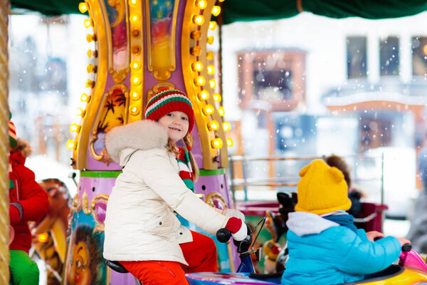 Kids at Christmas fair. Child at traditional street Xmas market in Germany. Winter outdoor fun. Little girl in knitted hat riding carousel horse in outdoor amusement park in winter holiday season. 