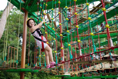 Child in forest adventure park. Kids climb on high rope trail. Agility and climbing outdoor amusement center for children. Little girl playing outdoors. School yard playground with rope way. clipart