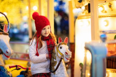 Çocuklar Noel Fuarı. Almanya geleneksel sokak Xmas pazarında çocuk. Kış açık eğlence. Kışın açık eğlence parkı içinde atlıkarınca at binme örgü şapkalı küçük kız tatil sezonu. 