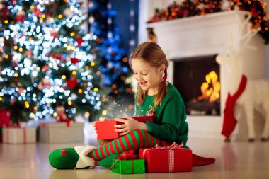 Evdeki Noel ağacında çocuk açılışı hediyesi. Noel hediyeleri ve oyuncakları olan elf kostümlü bir çocuk. Elinde hediye kutusu ve şöminede şekerlemesi olan küçük bir kız. Aile kış tatilini kutluyor. Ev dekorasyonu.