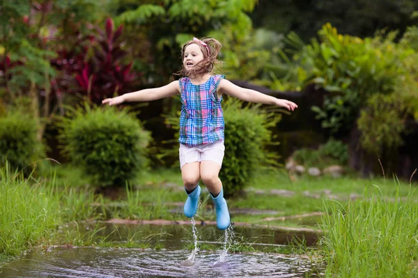 雨の中で遊んでいる子供 傘やレインブーツを着た子供たちは大雨の中屋外で遊ぶ 泥だらけの水たまりに飛び込む少年 子供たちは雨の秋の天気で楽しみます 熱帯低気圧の中を走る子供 — ストック写真