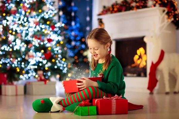 自宅のクリスマスツリーで子供のオープニングプレゼント クリスマスプレゼントやおもちゃで自分の衣装の子供 暖炉でギフトボックスとキャンディーを持つ小さな女の子 冬休みを祝う家族 家の装飾 — ストック写真