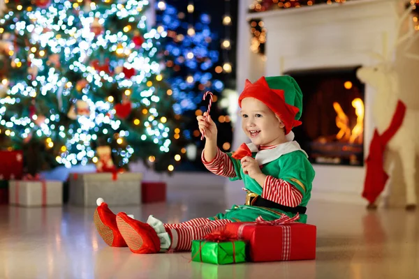 自宅のクリスマスツリーで子供のオープニングプレゼント クリスマスプレゼントやおもちゃで自分の衣装の子供 暖炉でギフトボックスとキャンディーを持つ小さな赤ちゃんの男の子 冬休みを祝う家族 家の装飾 — ストック写真