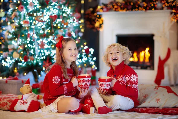 自宅にクリスマス ツリーの下の子 男の子とクリスマス飾り付きニット セーターの女の子は ホット チョコレートを飲みます 子供を持つ家族は 冬の休日を祝うため 子供たちは暖炉のそばでプレゼントを開けてください — ストック写真