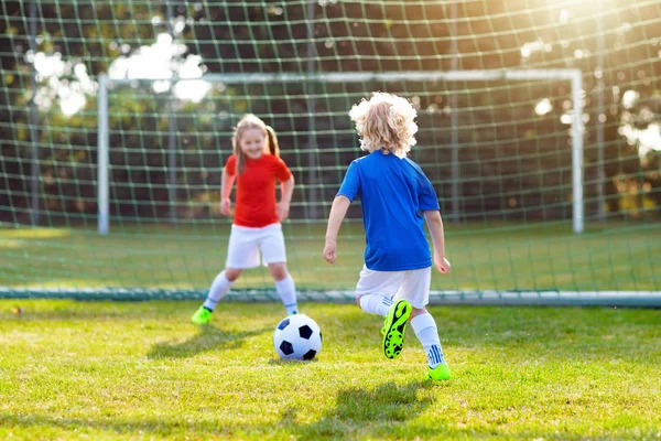 Kids play football on outdoor field. Children score a goal at soccer game. Girl and boy kicking ball. Running child in team jersey and cleats. School football club. Sports training for young player.