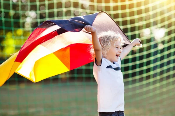 Kids play football on outdoor field. Germany team fans with national flag. Children score a goal at soccer game. Child in German jersey and cleats kicking ball. Fan celebrating victory at pitch.