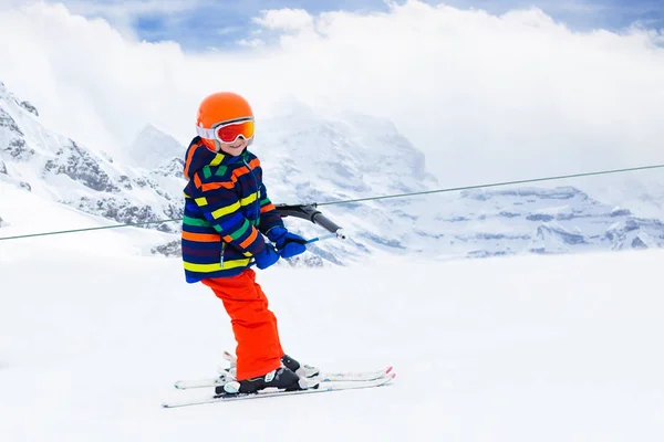 Child on a button ski lift going uphill in the mountains on a sunny snowy day. Kids in winter sport school in alpine resort. Family fun in the snow. Little skier learning and exercising on a slope.