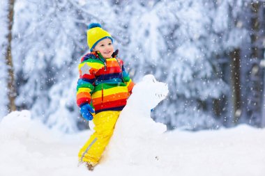 Karlı kış parkta kardan adam yapma çocuk. Çocuk karda oyun. Renkli ceket ve sonra kar yağışı kar dinozor kış bahçesinde bina şapka erkek bebek. Soğuk kış günü açık eğlence.