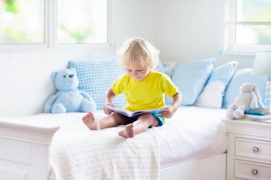 Beyaz güneşli bir odada pencereli bir çocuk yatakta oynuyor. Çocuk odası ve iç tasarım. Erkek bebek evde. Çocuk yuvası için yatak ve tekstil. Oyuncağı ve kitabı olan bir çocuk. Uyku ve uyku zamanı..