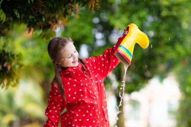 Sonbahar parkında yağmurda oynayan lastik çizmeli çocuk. Yağmurlu bir sonbahar gününde çamurlu su birikintisinde bir çocuk. Su dolu yağmur botu. Dışarıda, duşta kırmızı ceketli küçük bir kız. Çocuklar su geçirmez ayakkabılar.