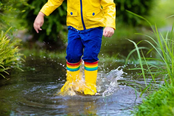 秋の公園で雨の中で遊んでいる子供 子供は雨の日に泥だらけの水たまりに飛び込む 激しいシャワーで雨のブーツと黄色のジャケットの屋外の小さな男の子 子供防水履物とコート — ストック写真