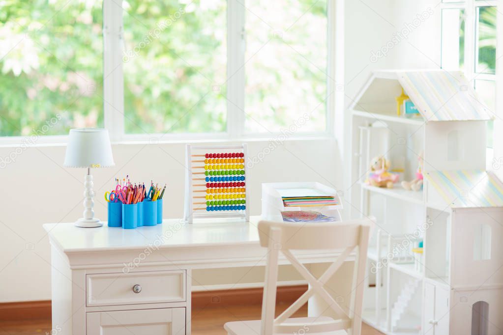 Kids bedroom with wooden desk and doll house. White sunny room with big window for young child. Home interior for little girl. Table for homework and study with lamp and abacus, books for school kid.