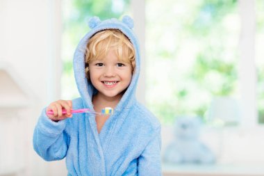 Child brushing teeth. Kids tooth brush and paste. Little baby boy in blue bath robe or towel brushing his teeth in white bathroom with window on sunny morning. Dental hygiene and heath for children. clipart