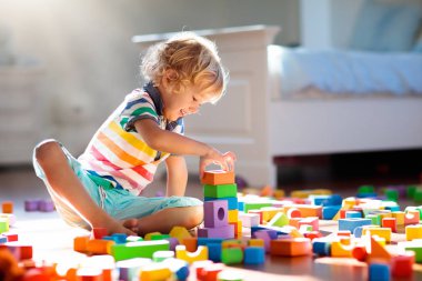 Çocuk renkli oyuncak bloklarıyla oynuyor. Çocuklar oynar. Küçük çocuk güneşli beyaz yatak odasındaki karanlık zeminde oyuncaklardan kule inşa ediyor. Bebek ve bebek için eğitici bir oyun. Çocuklar oyuncak ev yapar.