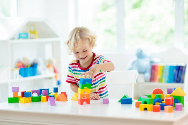 Kid playing with colorful toy blocks. Little boy building tower of block toys. Educational and creative toys and games for young children. Baby in white bedroom with rainbow bricks. Child at home.