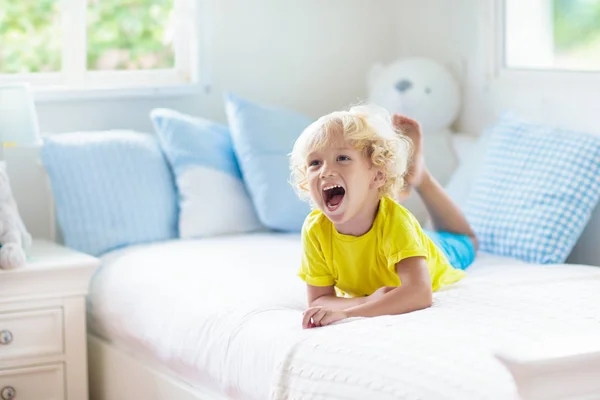 子供は窓付きの白い日当たりの良いベッドルームでベッドで遊ぶ キッズルームとインテリアデザイン 家にいる赤ん坊 子供の保育園のための寝具と繊維 おもちゃと本を持つ子供 昼寝と睡眠時間 — ストック写真