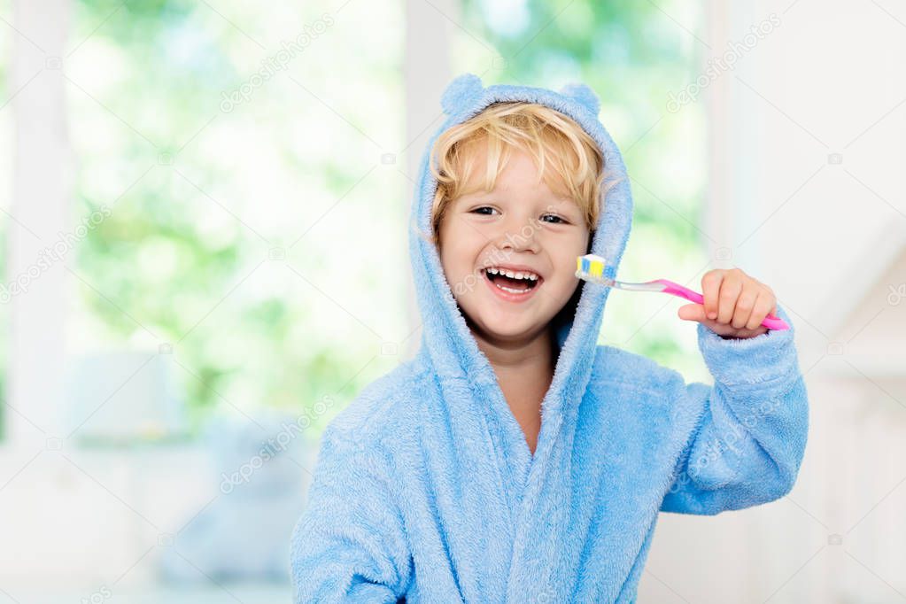Child brushing teeth. Kids tooth brush and paste. Little baby boy in blue bath robe or towel brushing his teeth in white bathroom with window on sunny morning. Dental hygiene and heath for children.