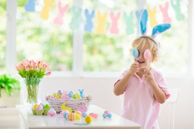 Çocuklar paskalya yumurtalarını boyuyor. Tavşan kulaklı çocuklar Paskalya avı için renkli yumurta boyuyorlar. Bahar tatili kutlaması için çiçek, sepet ve tavşanlı ev dekorasyonu. Küçük bebek evi dekore ediyor..