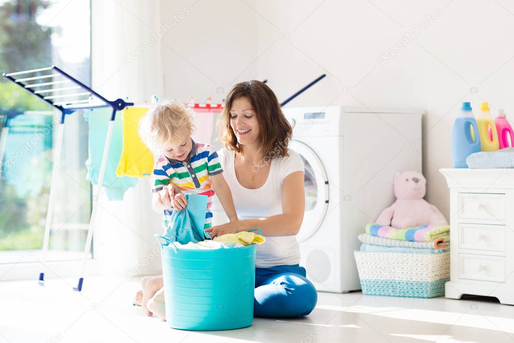 Mother and kids in laundry room with washing machine or tumble dryer. Family chores. Modern household devices and washing detergent in white sunny home. Clean washed clothes on drying rack. 