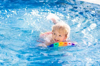 Çocuk tropikal tatil köyü havuzunda yüzmeyi öğreniyor. Çocuklar yüzmeyi öğrenir. Küçük çocuklar için egzersiz ve eğitim. Spor kulübünde renkli sörf tahtası olan küçük bir çocuk. Yüzen bebek ya da bebek.
