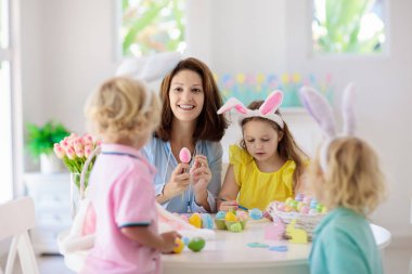 Anne ve çocuklar Paskalya yumurtalarını boyar. Anne, tavşan kulaklı küçük kız ve oğlan ölüyor ve beyaz güneşli odada Paskalya yumurtası avı için resim yapıyorlar. Aile kutlaması ve bahar tatili için ev dekorasyonu..