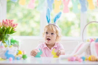 Çocuklar paskalya yumurtalarını boyuyor. Tavşan kulaklı çocuklar Paskalya avı için renkli yumurta boyuyorlar. Bahar tatili kutlaması için çiçek, sepet ve tavşanlı ev dekorasyonu. Küçük bebek evi dekore ediyor..