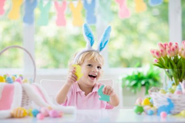 Çocuklar paskalya yumurtalarını boyuyor. Tavşan kulaklı çocuklar Paskalya avı için renkli yumurta boyuyorlar. Bahar tatili kutlaması için çiçek, sepet ve tavşanlı ev dekorasyonu. Küçük bebek evi dekore ediyor..