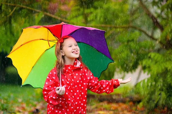 秋の公園で雨の中で遊んでいる子供 傘とレインブーツを持つ子供は大雨の中で屋外で遊ぶ 秋のシャワーの下で赤いジャケットの少女 雨で子供たちは楽しい 嵐の中で遊ぶ子供たち — ストック写真