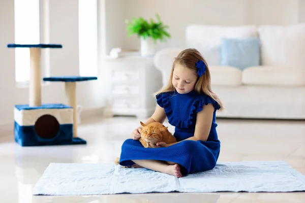 Child playing with cat at home. Kids and pets. Little girl feeding and petting cute ginger color cat. Cats tree and scratcher in living room interior. Children play and feed kitten. Home animals.