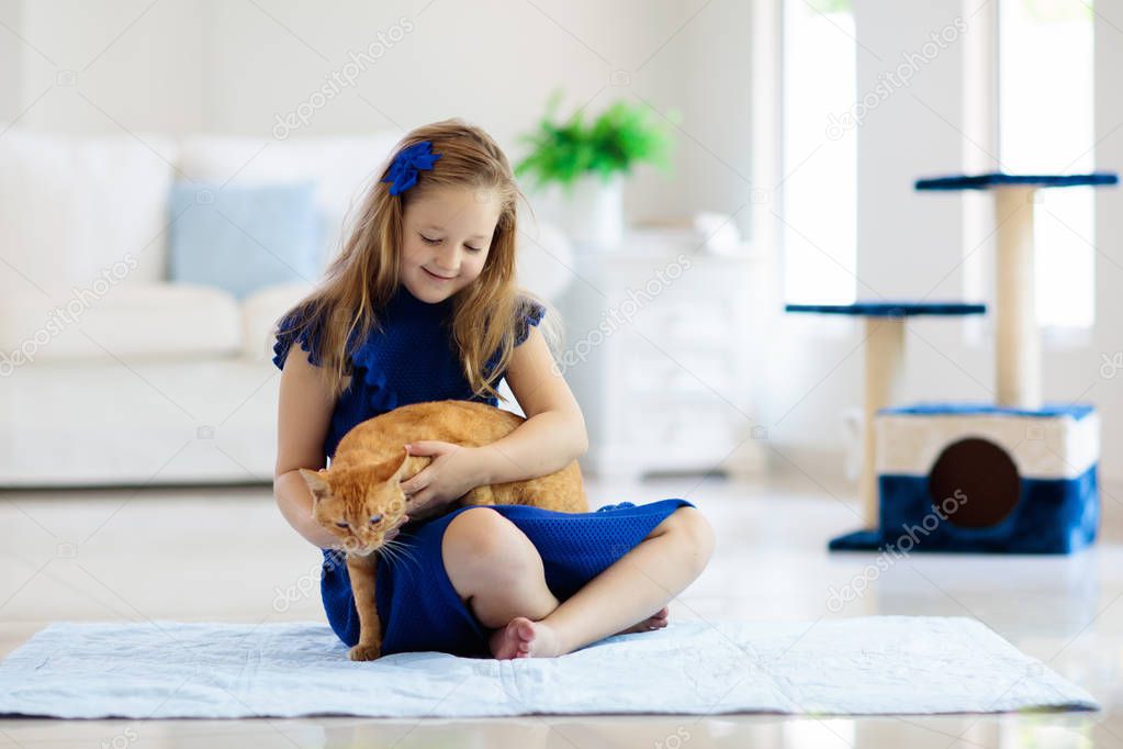 Child playing with cat at home. Kids and pets. Little girl feeding and petting cute ginger color cat. Cats tree and scratcher in living room interior. Children play and feed kitten. Home animals.