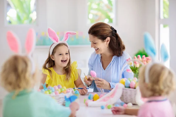 Mother and kids color Easter eggs. Mom, little girl and boy with bunny ears dying and painting for Easter egg hunt in white sunny room. Family celebration and home decoration for spring holiday.