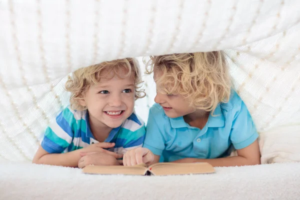 Child reading book in bed under knitted blanket. Two brothers playing together. Kids cozy bedroom hygge style. Little boy doing homework before sleep.