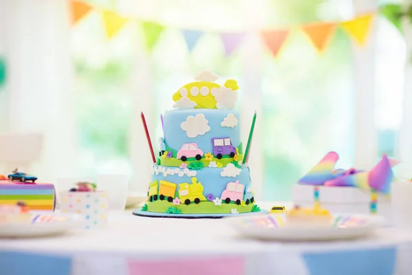 Kids birthday party. Boy cake with car and airplane. Child blowing out candles on colorful cake. Party decorations, rainbow flag banners, balloons. Vehicle theme celebration. Kid celebrating birthday