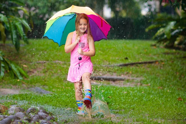 雨の中で遊んでいる子供 傘やレインブーツを着た子供たちは大雨の中屋外で遊ぶ 泥だらけの水たまりに飛び込む少女 子供たちは雨の秋の天気で楽しみます 熱帯低気圧の中を走る子供 — ストック写真