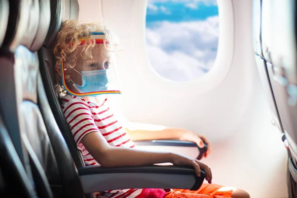 Child in airplane in face shield and mask. Flying with kids during virus outbreak. Vacation in coronavirus and flu pandemic. Safe travel with young child and baby. Kids fly airplane in surgical masks.